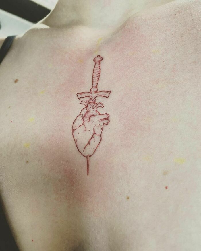 Red ink tattoo of a heart with a sword design on skin.