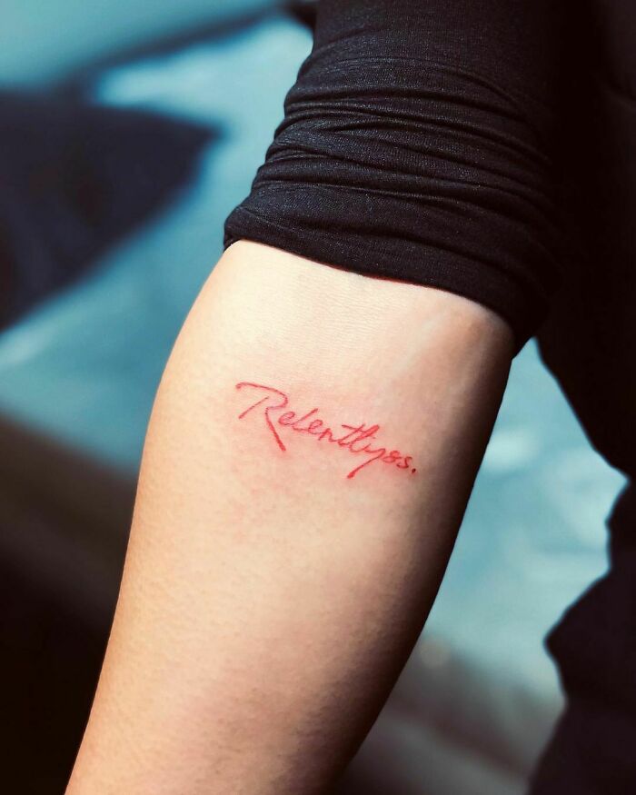 Arm with "Relentless" red ink tattoo, showcasing minimalist design style.
