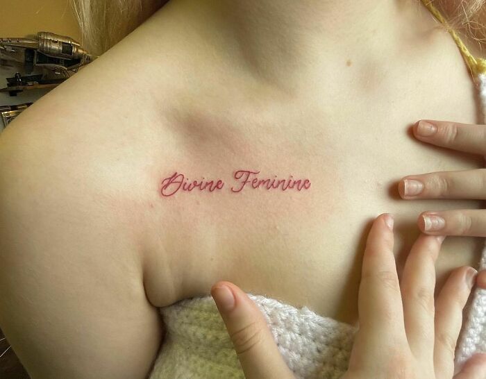 Red ink tattoo reading "Divine Feminine" on the collarbone.