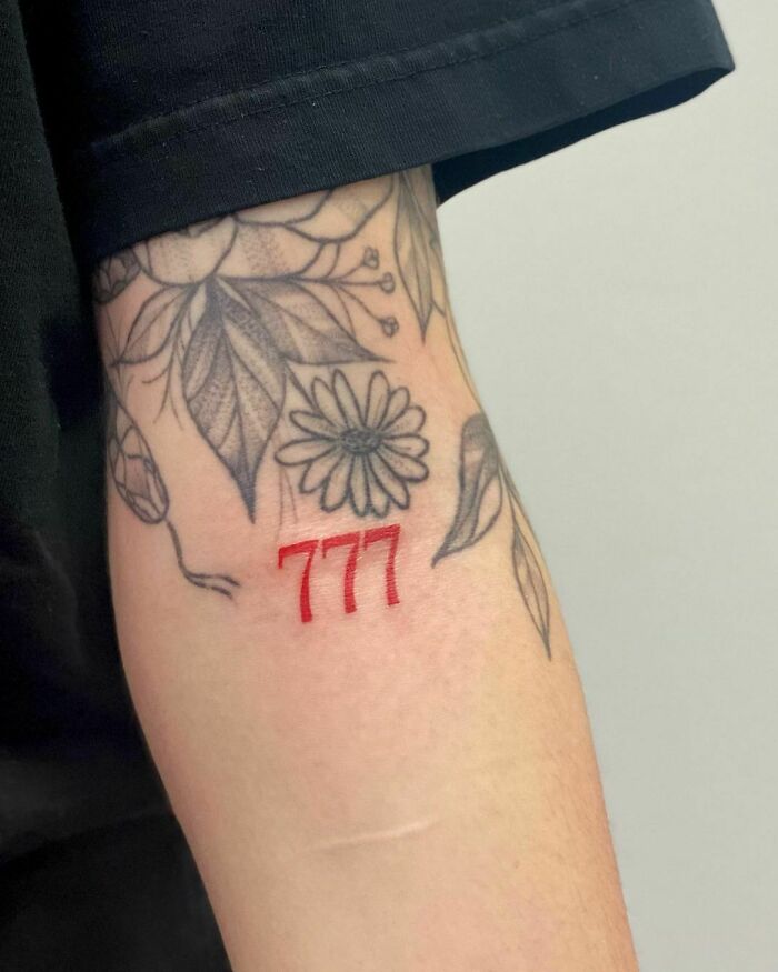 Red ink tattoo of "777" on an arm with floral black ink designs around it.