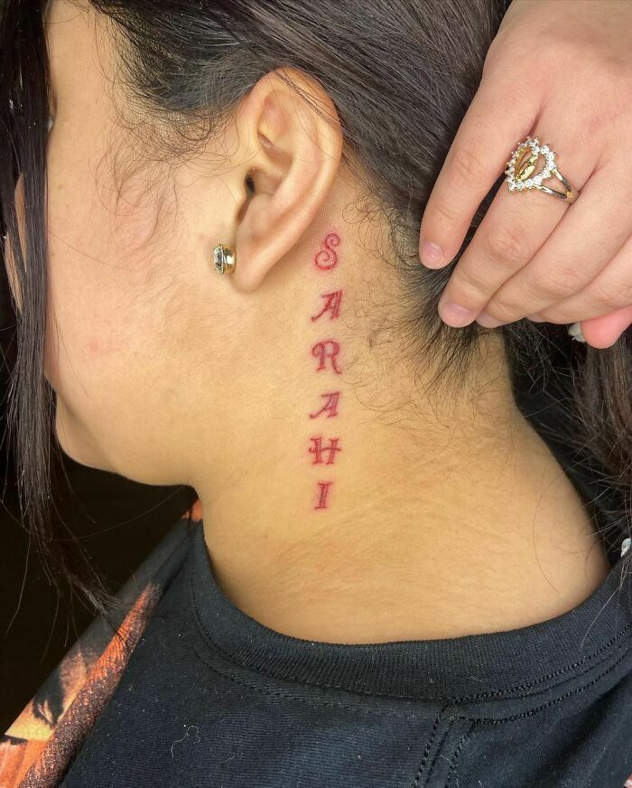 Red ink tattoo design behind ear, spelling "SARAHI" in elegant script.