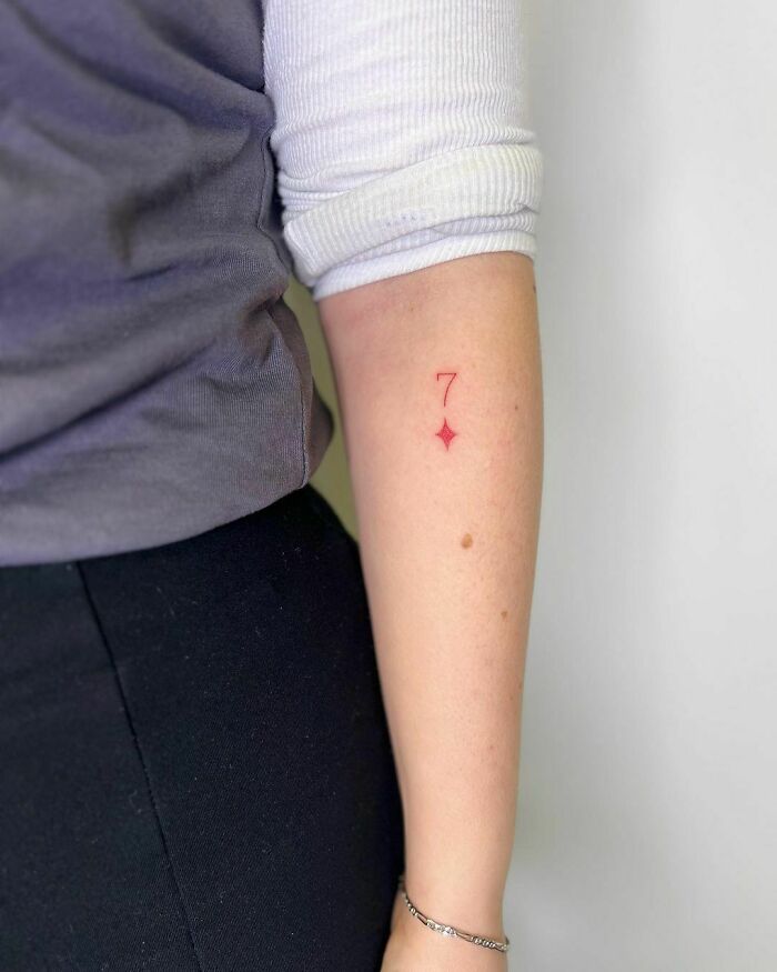 Red ink tattoo of the number 7 with a star on a person's forearm.
