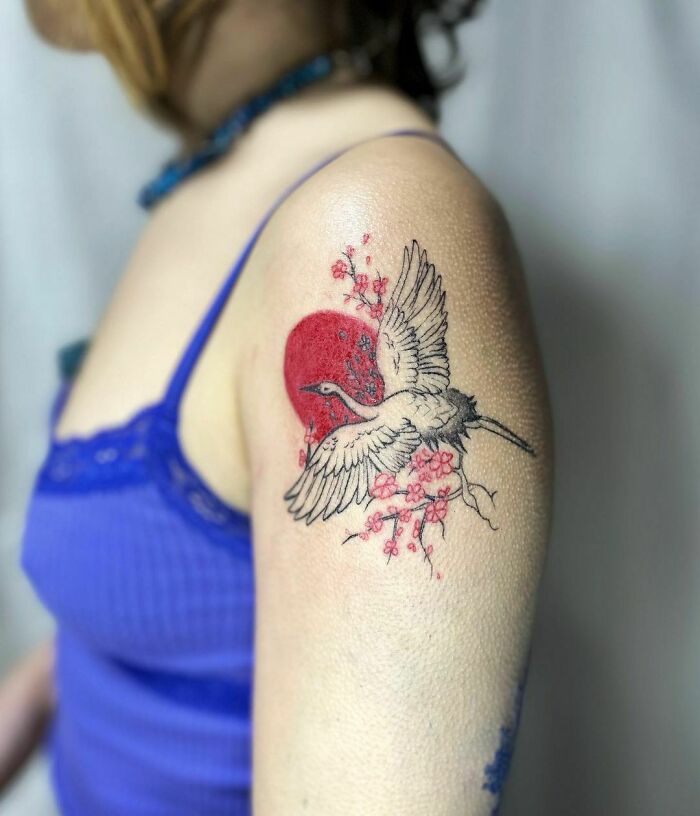 Red ink tattoo of a crane with cherry blossoms and a sun on a person's upper arm.