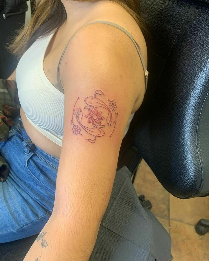 Woman with a floral design in red ink tattoo on her upper arm, seated in a chair.
