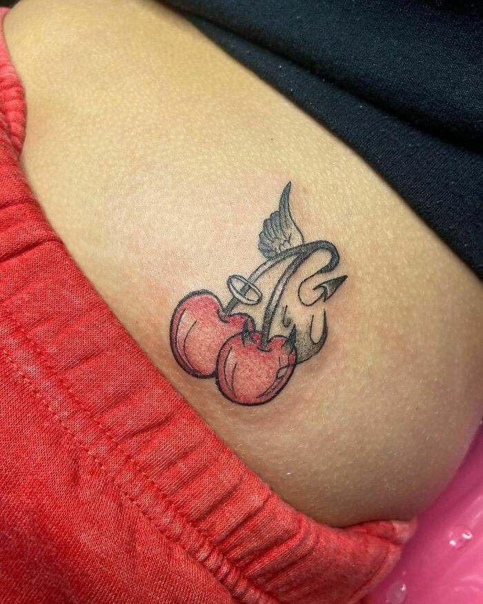 Red ink tattoo design of cherries with a wing and devil tail on skin.