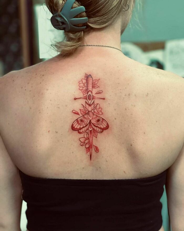 A person with a decorative red ink tattoo of a dagger and moth on their upper back.