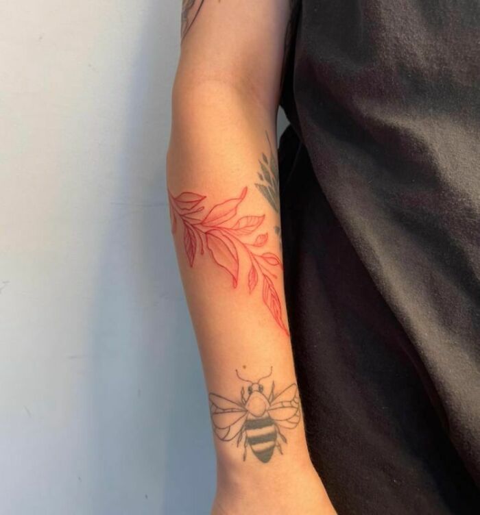 A forearm with a red ink tattoo featuring a leaf design and a black bee tattoo below.