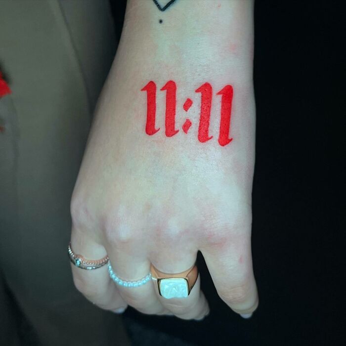 Red ink tattoo of "11:11" on a hand, featuring rings and minimalist design.