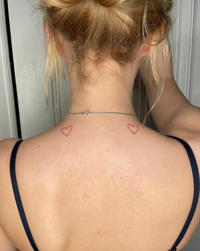 Back view of two small red ink heart tattoos on the woman's upper back.