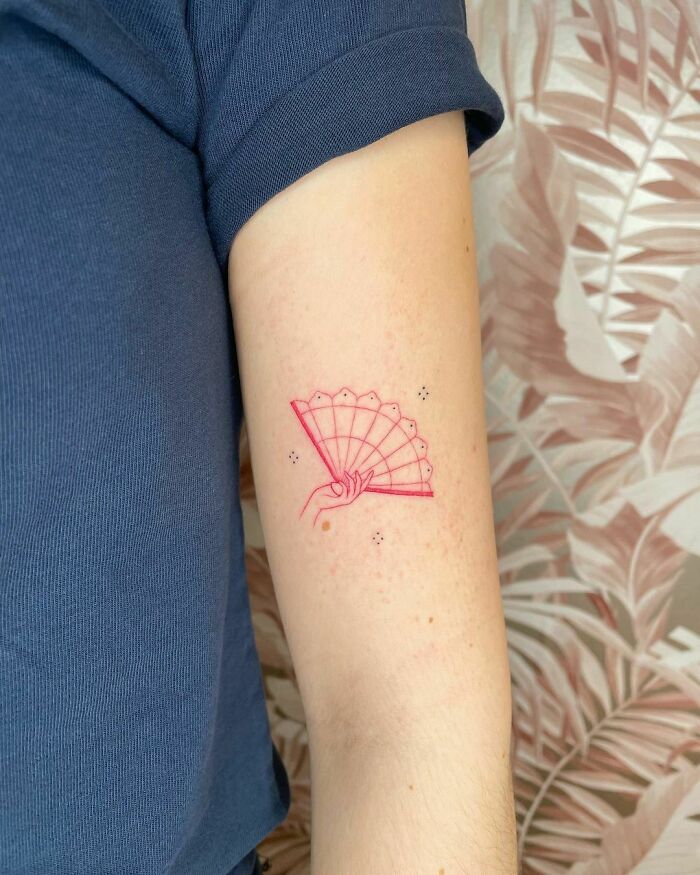 Red ink tattoo of a hand holding a fan on an arm, set against a patterned background.
