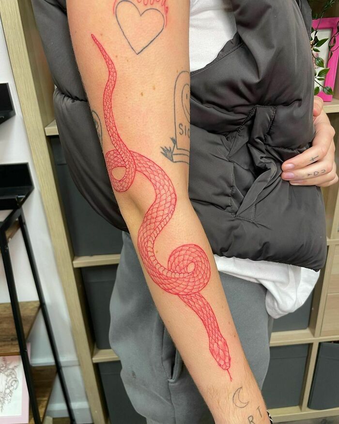 Red ink tattoo of a snake on an arm with various other small tattoos visible in the background.