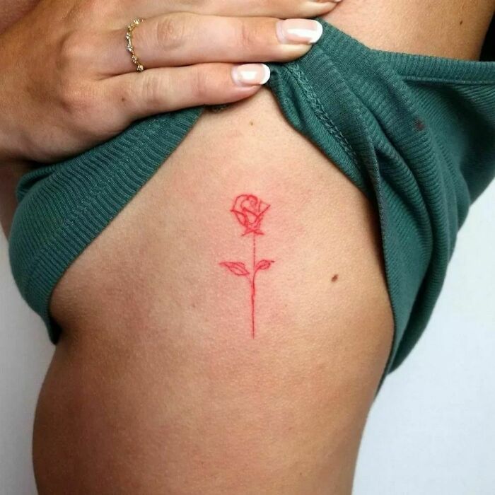 Red ink tattoo of a rose on someone's side, showcased by lifting a green shirt.
