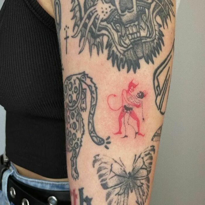Red ink tattoo of a small devil design on an arm, amidst other black ink tattoos.