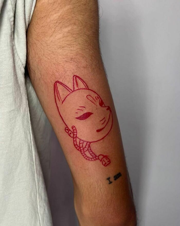 Red ink tattoo of a cat face on an arm, featuring simple line art and a rope detail.