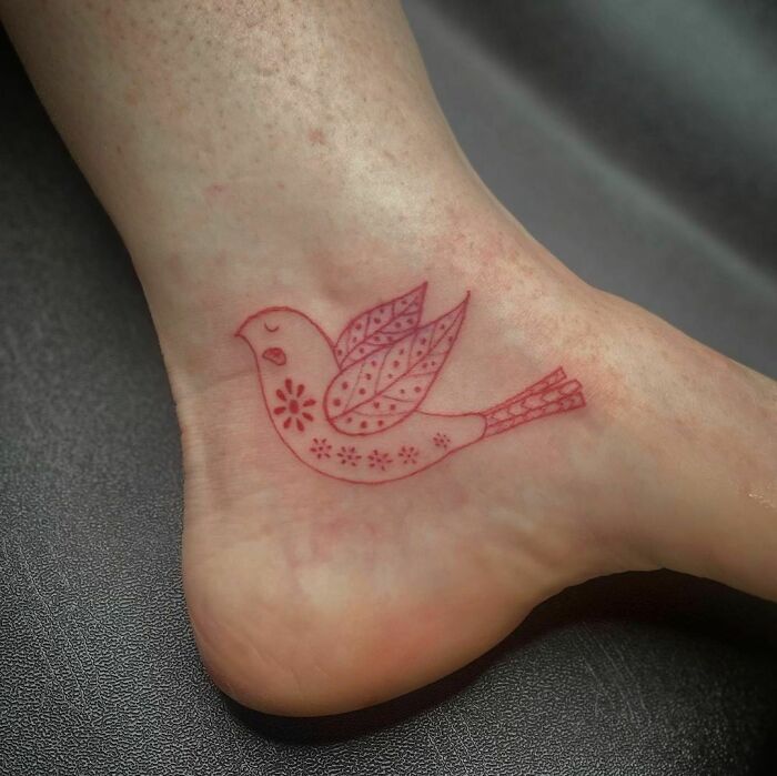Red ink tattoo of a stylized bird on an ankle, showcasing intricate design and patterns.