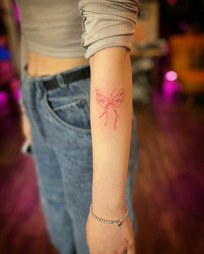A delicate red ink butterfly tattoo on a person's forearm, wearing a casual outfit, showcasing red tattoo design.