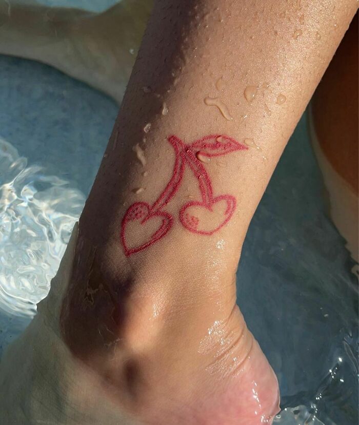 Red ink tattoo of heart-shaped cherries on ankle, with droplets of water.