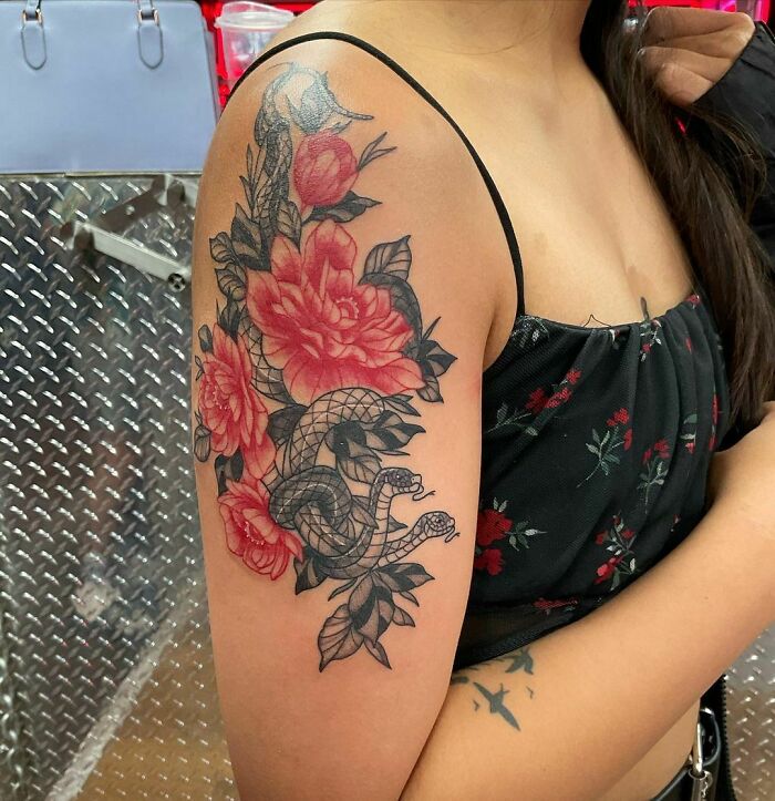 Woman with a detailed red ink tattoo of flowers and a snake on her upper arm, wearing a black floral top.