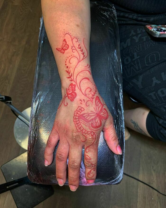 Red ink tattoo featuring butterflies and swirls on a hand, showcasing delicate and intricate design elements.