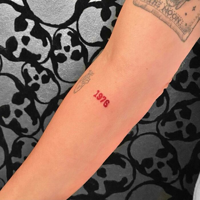 Red ink tattoo of "1976" on an arm with skull-pattern background.
