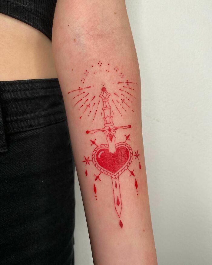 Red ink tattoo design featuring a dagger through a heart on forearm.