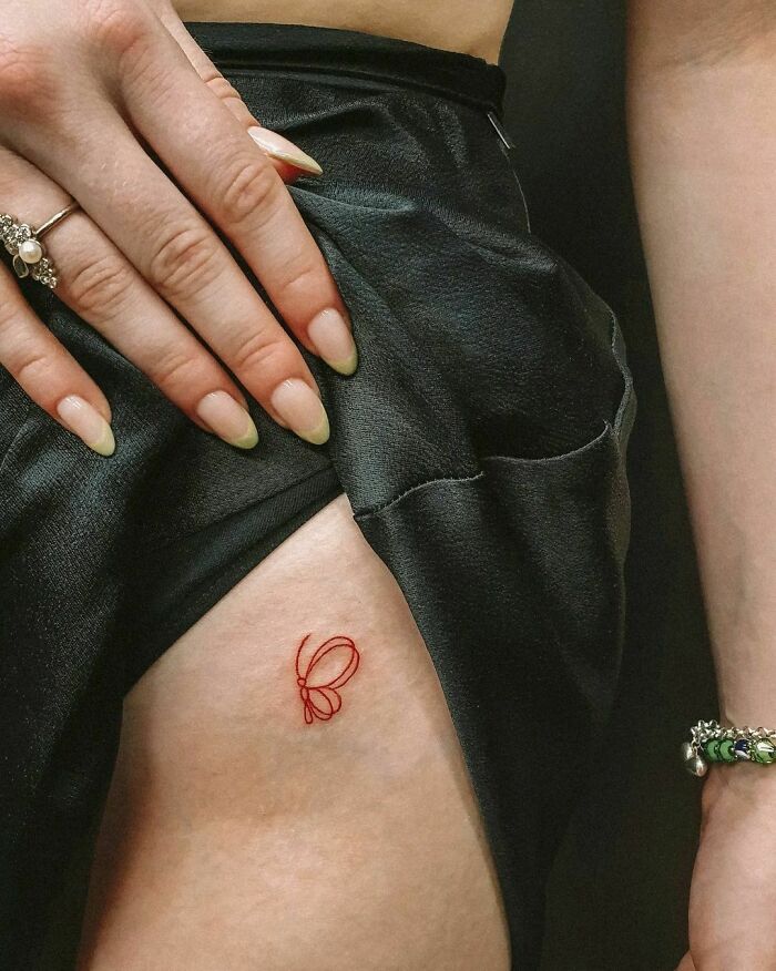 A small red ink tattoo of a butterfly on the thigh, partially covered by black fabric.