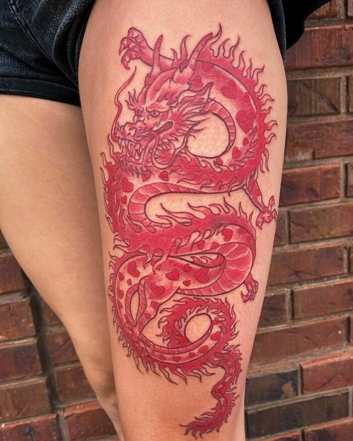 Red ink tattoo of a dragon on a person's thigh, showcasing intricate design details against a brick wall backdrop.
