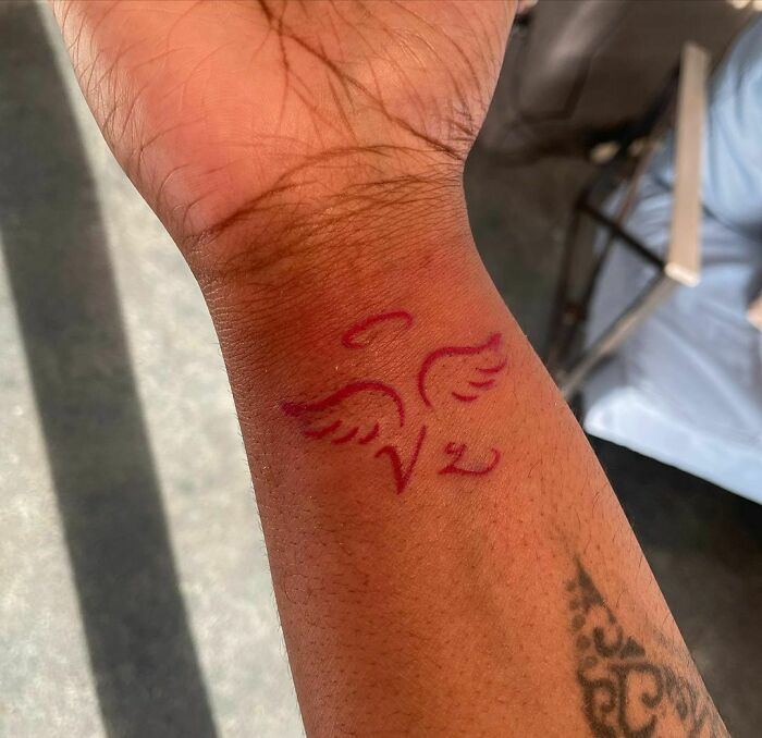 Red ink tattoo of wings and a v symbol on a person's wrist.