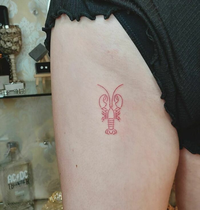 Red ink tattoo of a small lobster on the thigh, showcasing minimalist tattoo design.