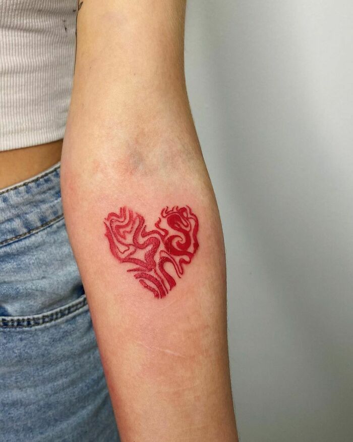 Red ink tattoo design on forearm, featuring an abstract heart shape with intricate patterns.