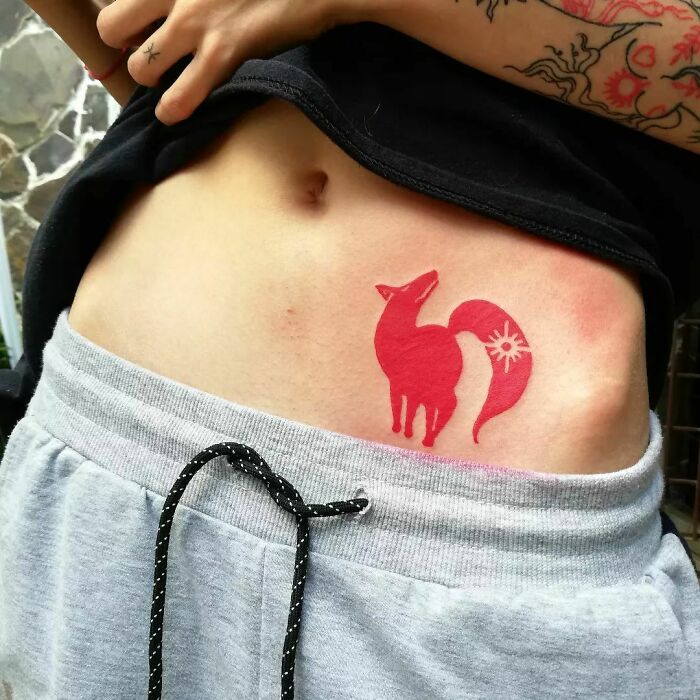 Red ink tattoo of a fox design on a person's side, showcasing vibrant red tattoo ideas in grayscale sweatpants.