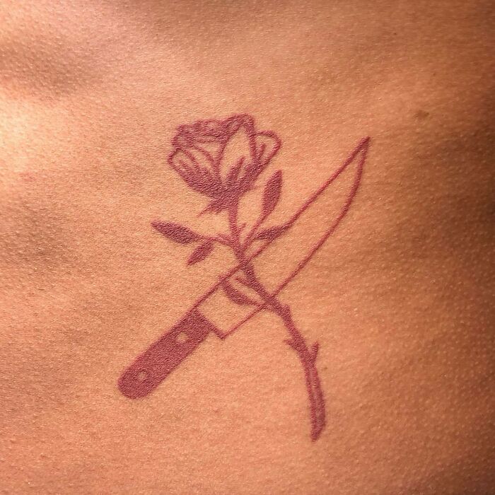 Red ink tattoo of a rose intertwined with a knife on skin.