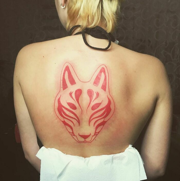 Red ink tattoo of a stylized fox face on a person's back with blonde hair, showcasing intricate design elements.
