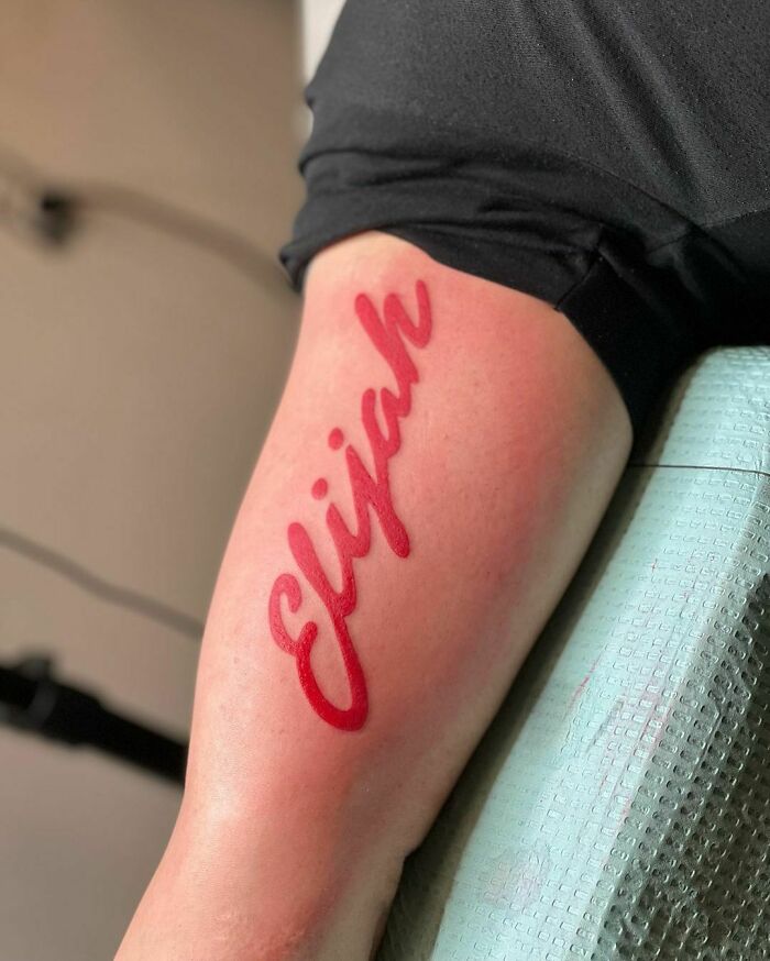 Red ink tattoo on arm with the name "Elijah" in cursive script, showcasing a bold design.