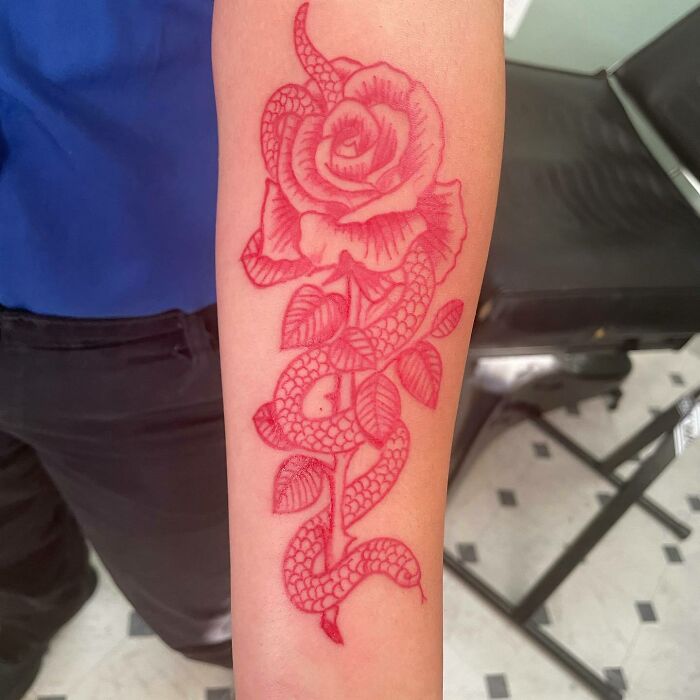Red ink tattoo of a rose entwined with a snake on a forearm.