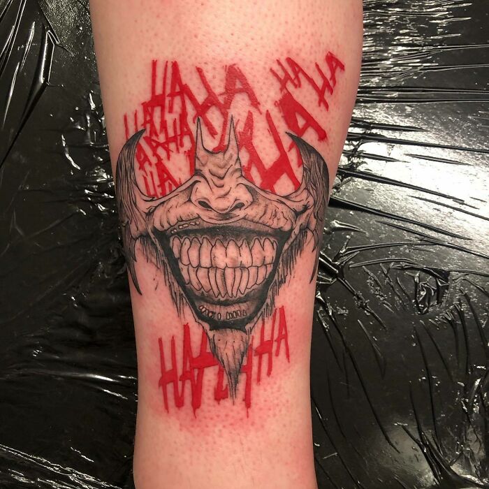 Red tattoo design of a grinning demon face with "HA HA" in bold red ink on forearm.