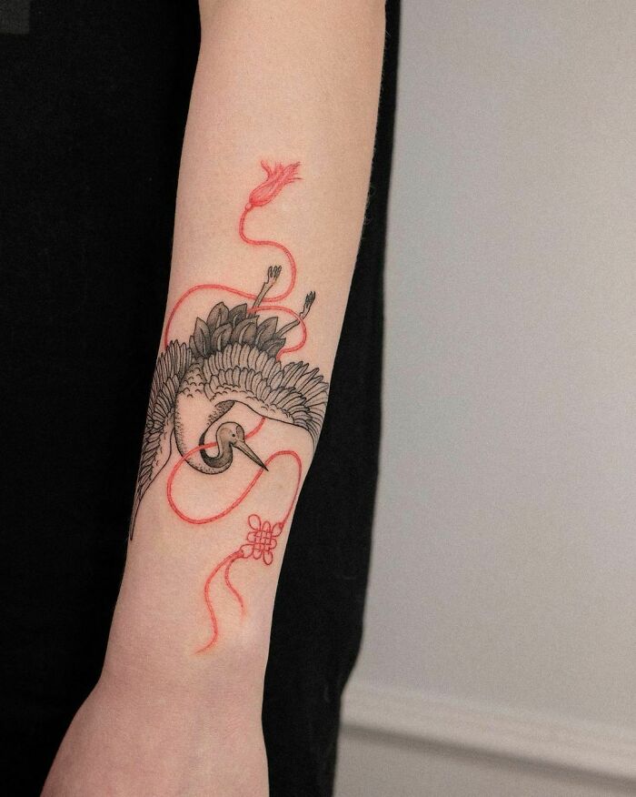 Red ink tattoo of a stylized crane and ribbon on forearm, showcasing intricate detail and bold design.