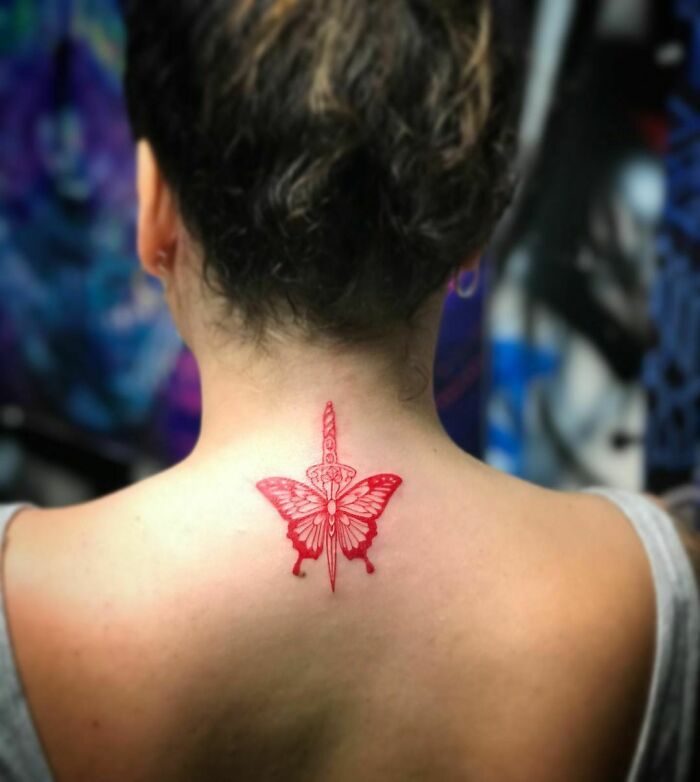 Red ink tattoo of a butterfly with a sword on a person's upper back.