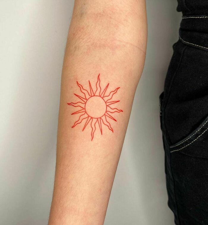 Red ink tattoo of a sun design on forearm, showcasing intricate linework.