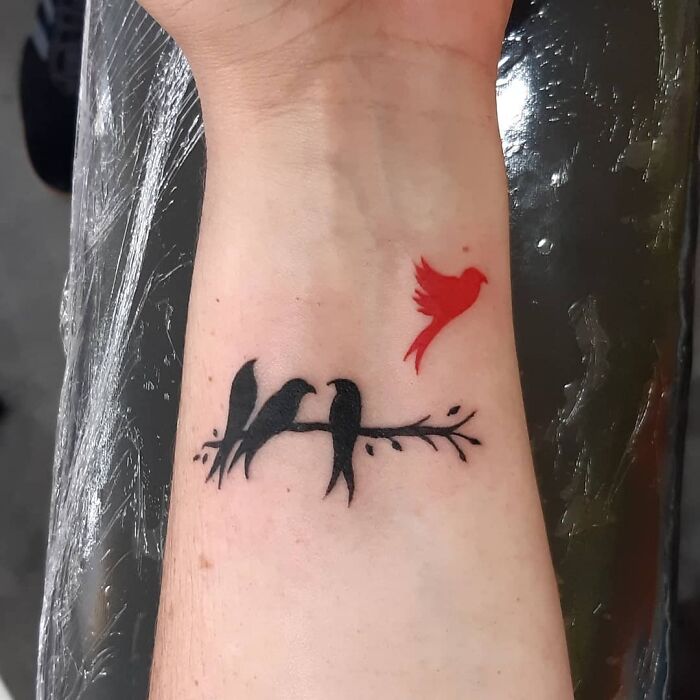 A black and red ink tattoo of birds on a branch with a flying red bird above on the forearm.