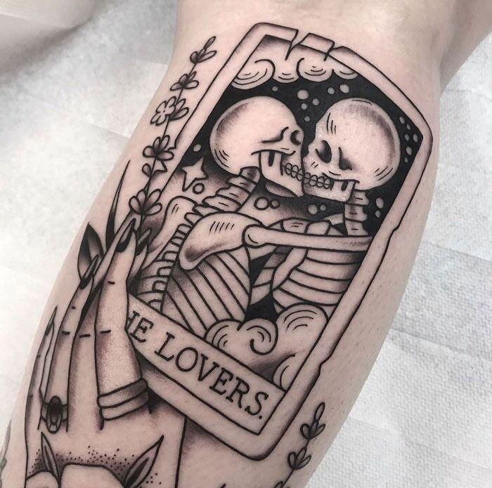 Lower leg tattoo featuring skeletons in an embrace within a tarot card labeled "The Lovers."