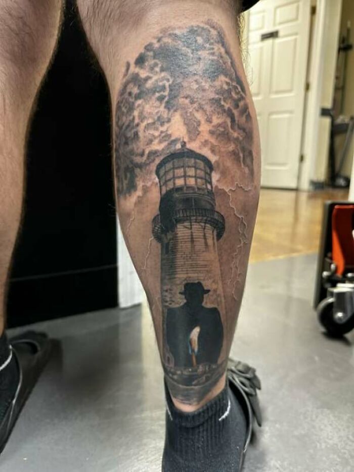 Lower leg tattoo featuring a lighthouse with a stormy sky and silhouette of a person in front.