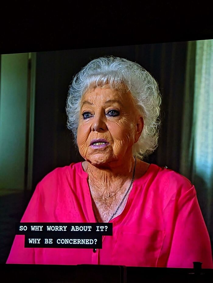 Elderly woman with exaggerated makeup, wearing a bright pink top, speaking with subtitles on screen.