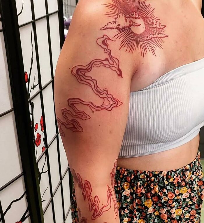 Woman with intricate red ink tattoo on upper arm featuring sun and clouds, wearing a white crop top.