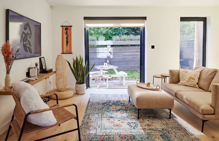 Cozy living room with modern home decoration, featuring a beige sofa, woven chair, and colorful rug near a garden view.
