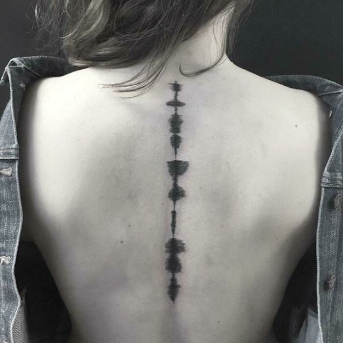 Elegant spine tattoo on a woman's back, featuring a symmetrical black design, with a denim jacket draped over her shoulders.