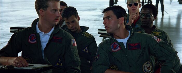 Two pilots in flight suits sitting together, representing an iconic best friend duo in a hangar setting.
