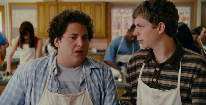 Two best friends in a kitchen scene, wearing aprons, engaged in a conversation.