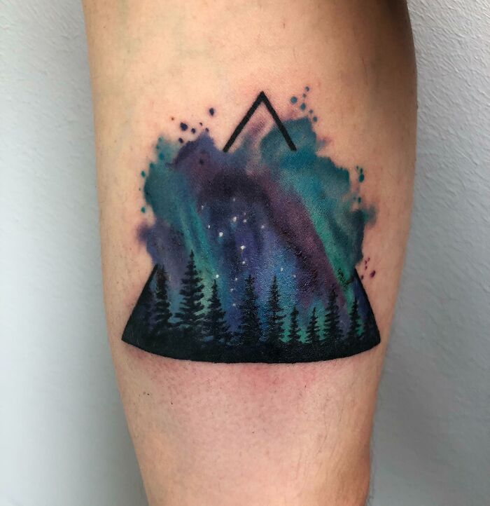 Tattoo of a colorful galaxy inside a silhouette of a mountain and forest, symbolizing a wild connection to nature.