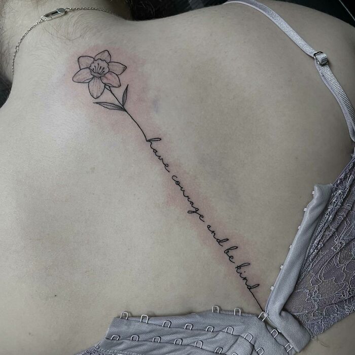 Delicate spine tattoo with a flower and cursive text "have courage and be kind" along the back.